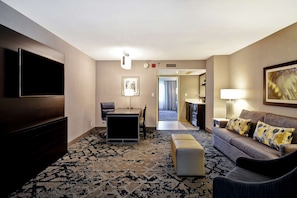 A hotel room with a flat-screen TV, a sofa with patterned cushions, a dining area with a table and chairs, and a patterned carpet.