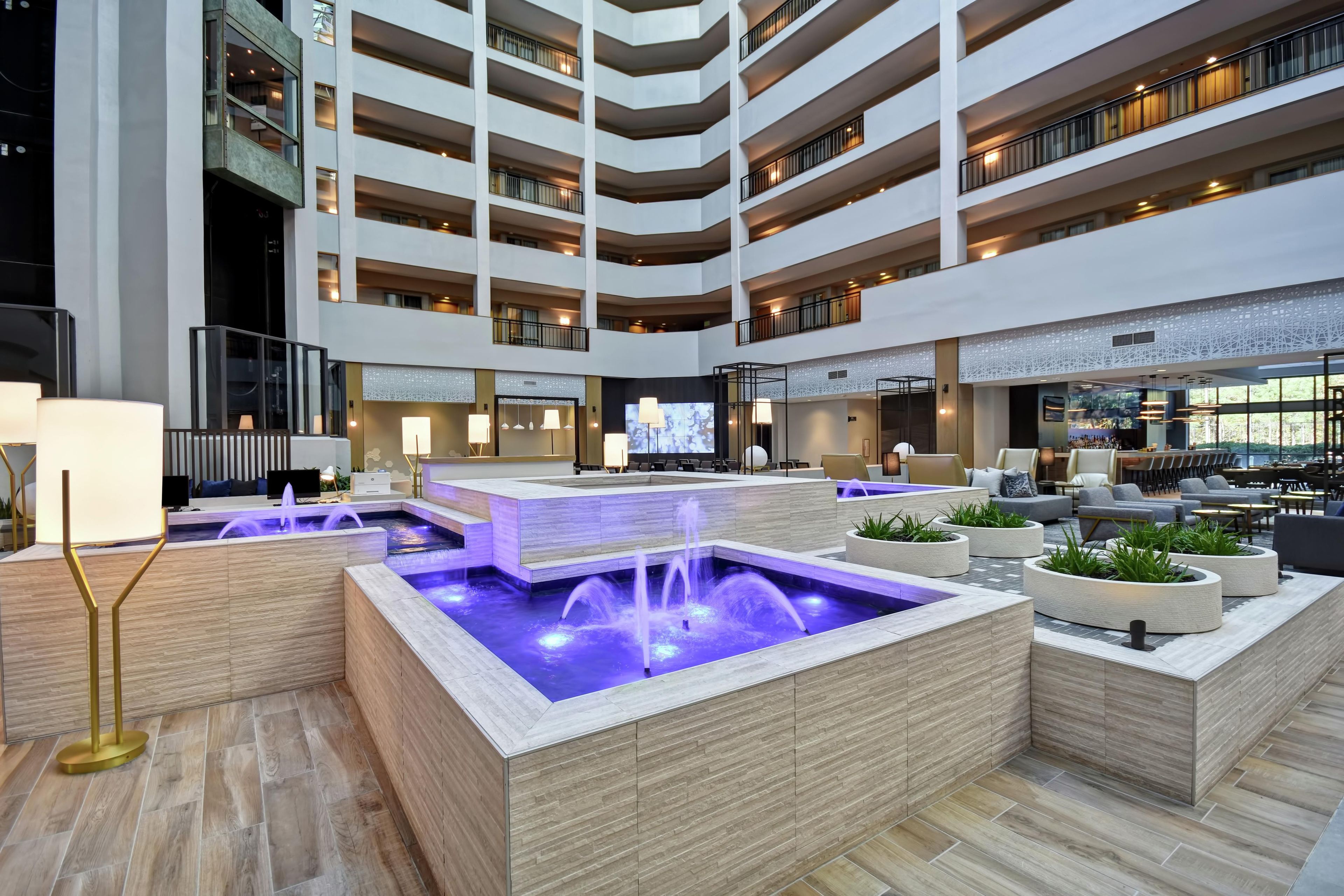 Un hall d’hôtel à plusieurs niveaux, doté d’un élément décoratif central en eau, d’espaces de détente et d’un bar.