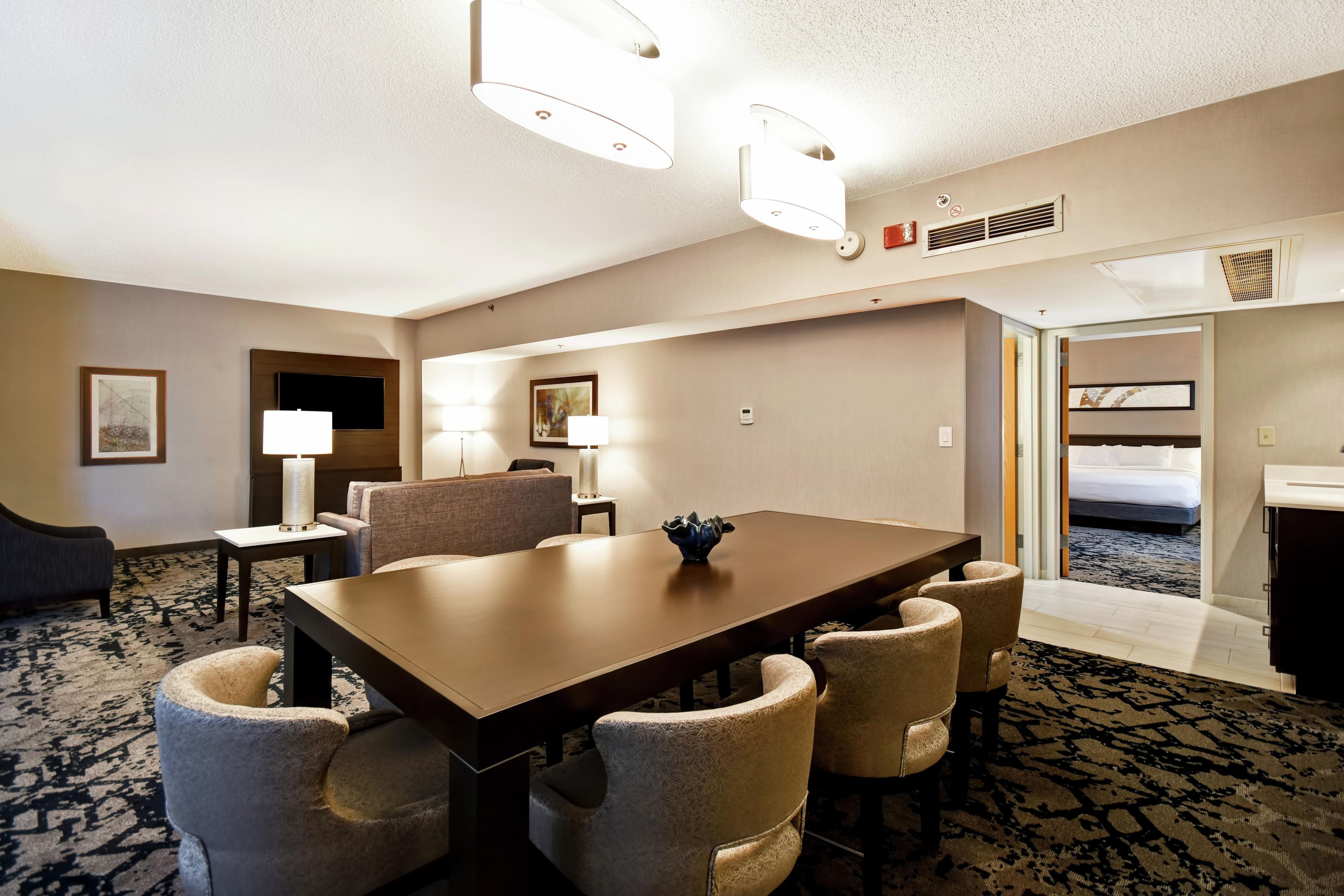 A hotel room with a large dining table, comfortable seating, a flat-screen TV, and a bed.