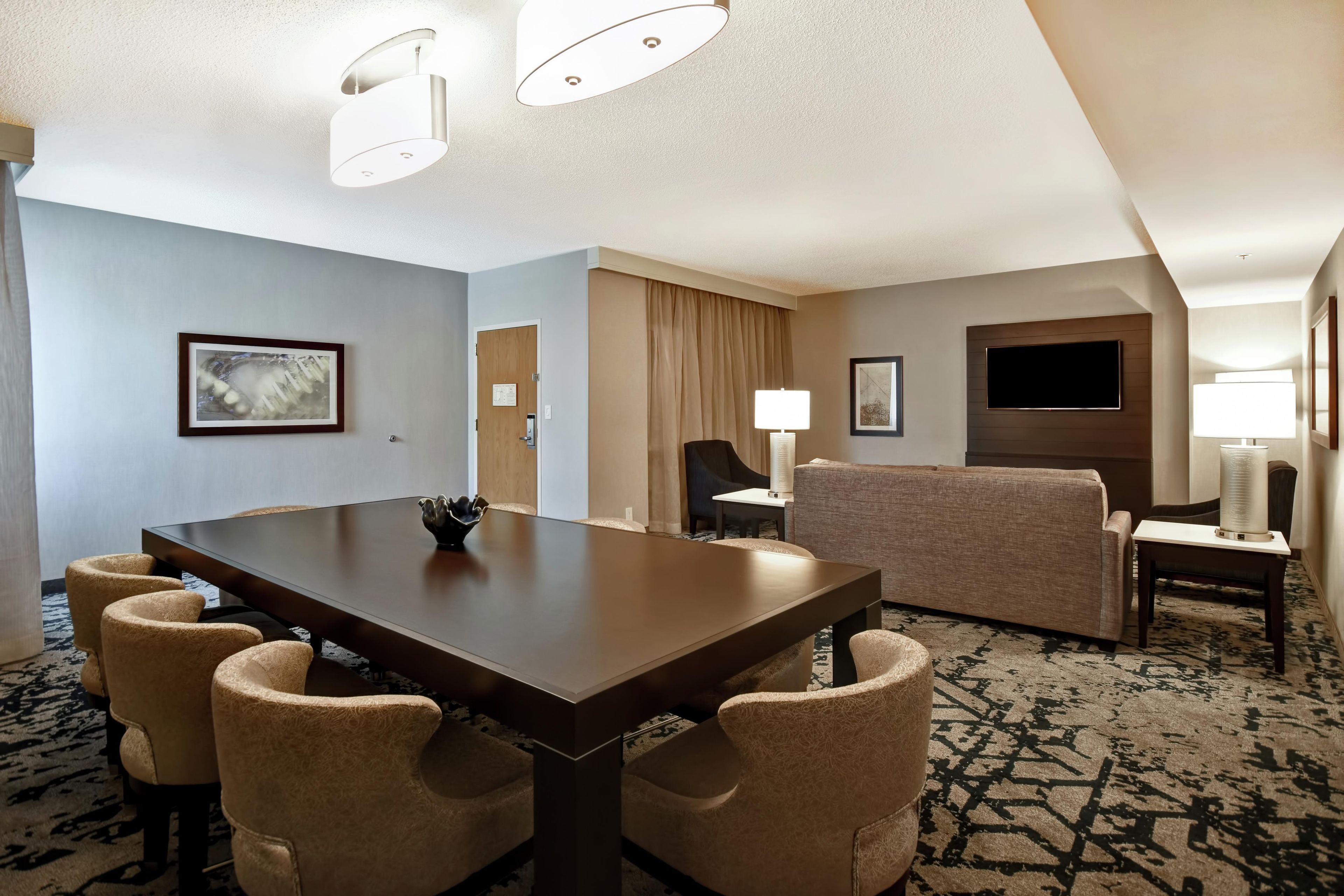 A hotel room with a dining table, four chairs, a sofa, a TV, and a lamp.
