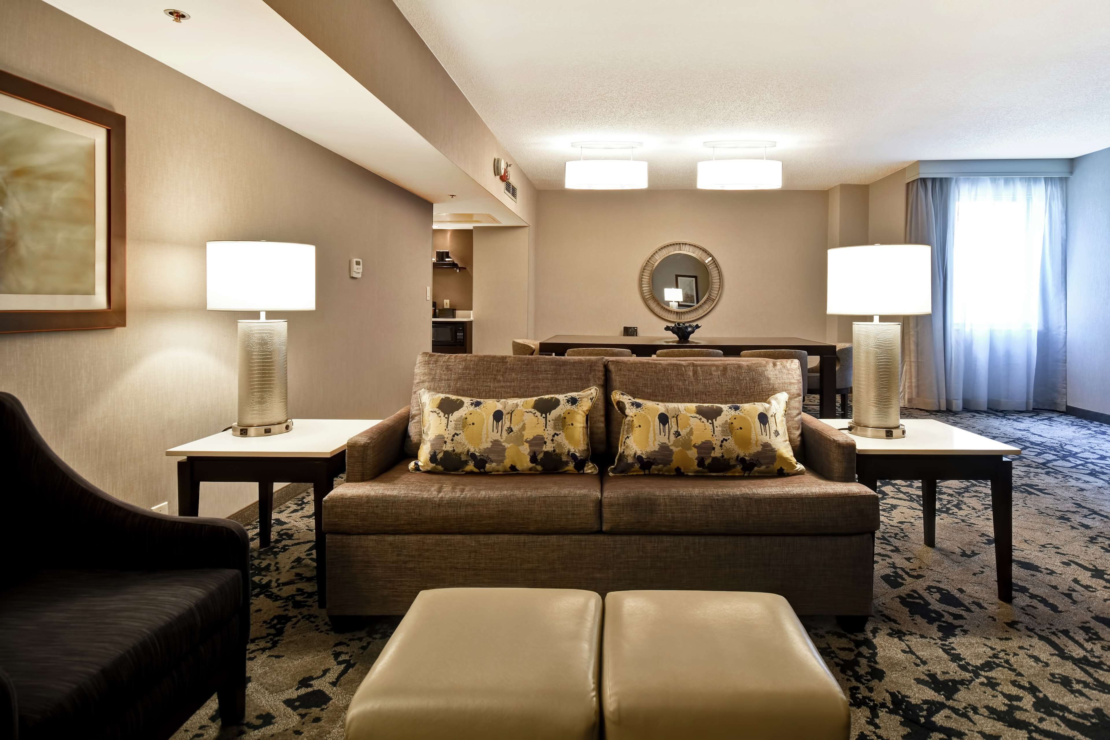 A hotel room with a sofa, two side tables with lamps, a coffee table, a dark armchair, a patterned carpet, and a large mirror on the wall.