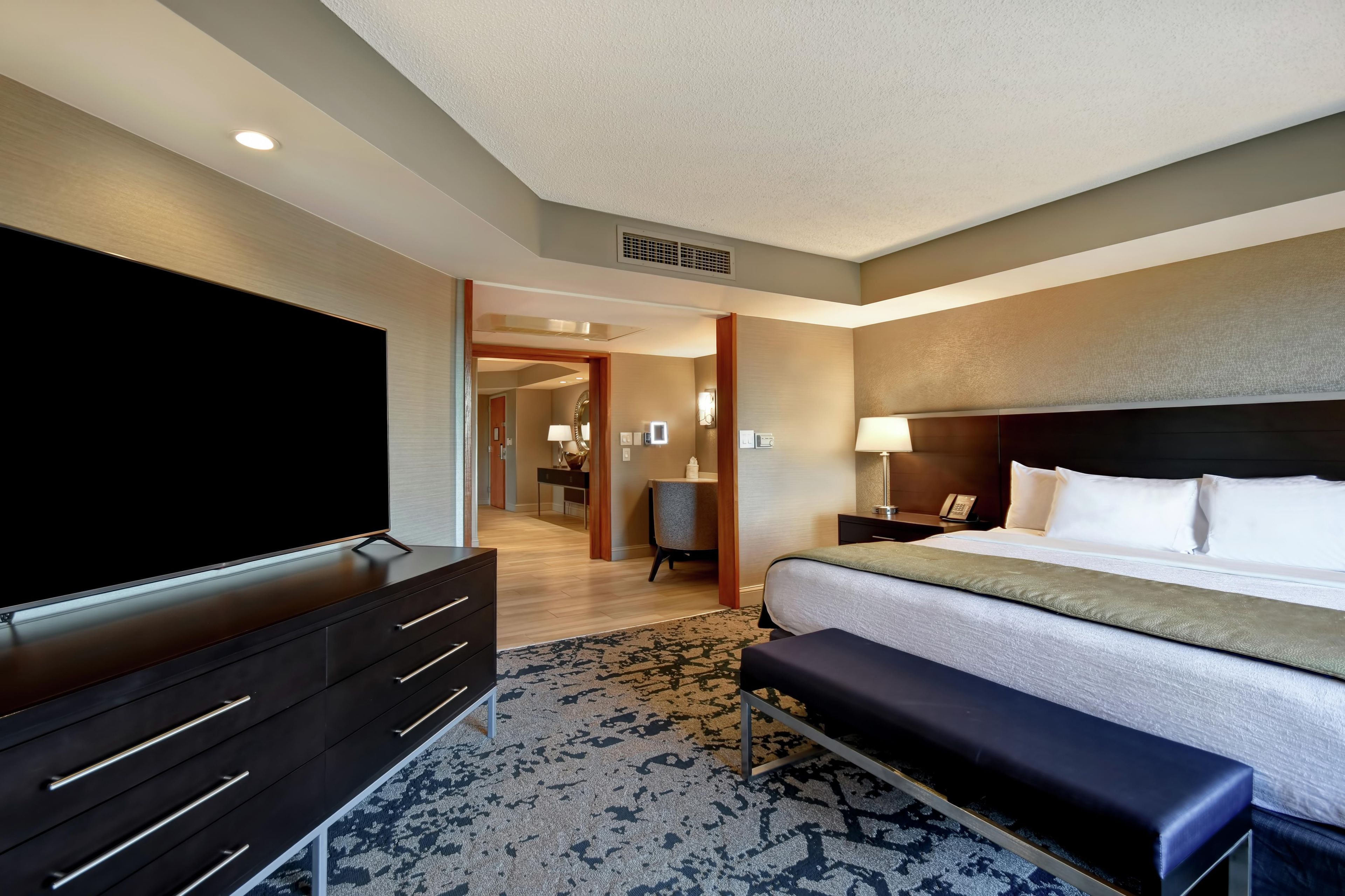 A hotel room with a large flat-screen TV, a bed with a headboard, a bench, a desk with a lamp, and a bathroom visible through an open door.