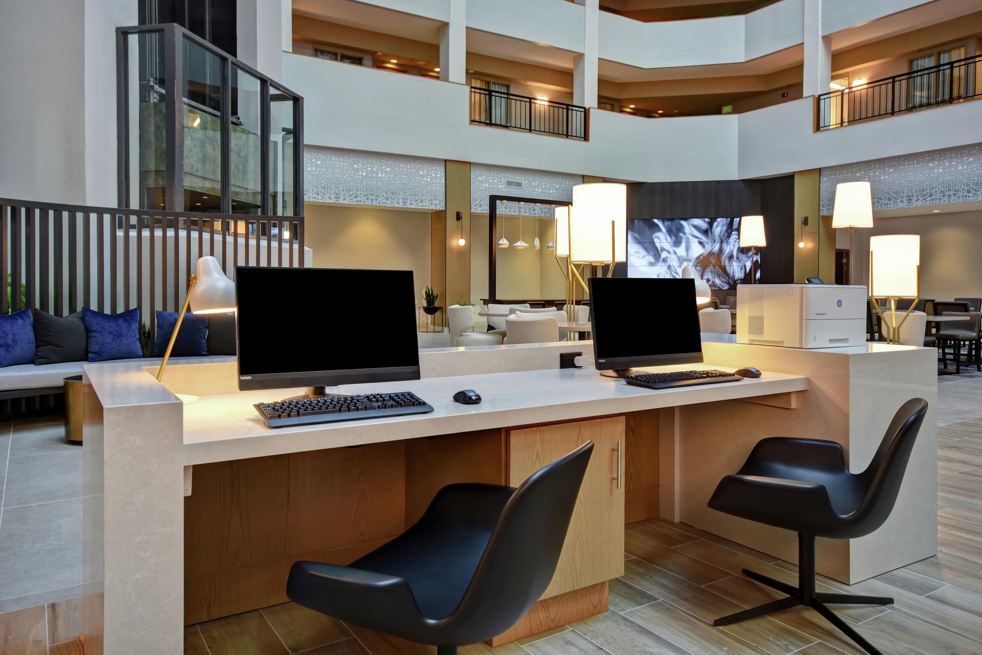 A modern hotel lobby with a central reception desk, two computer stations, and a seating area with sofas and chairs.