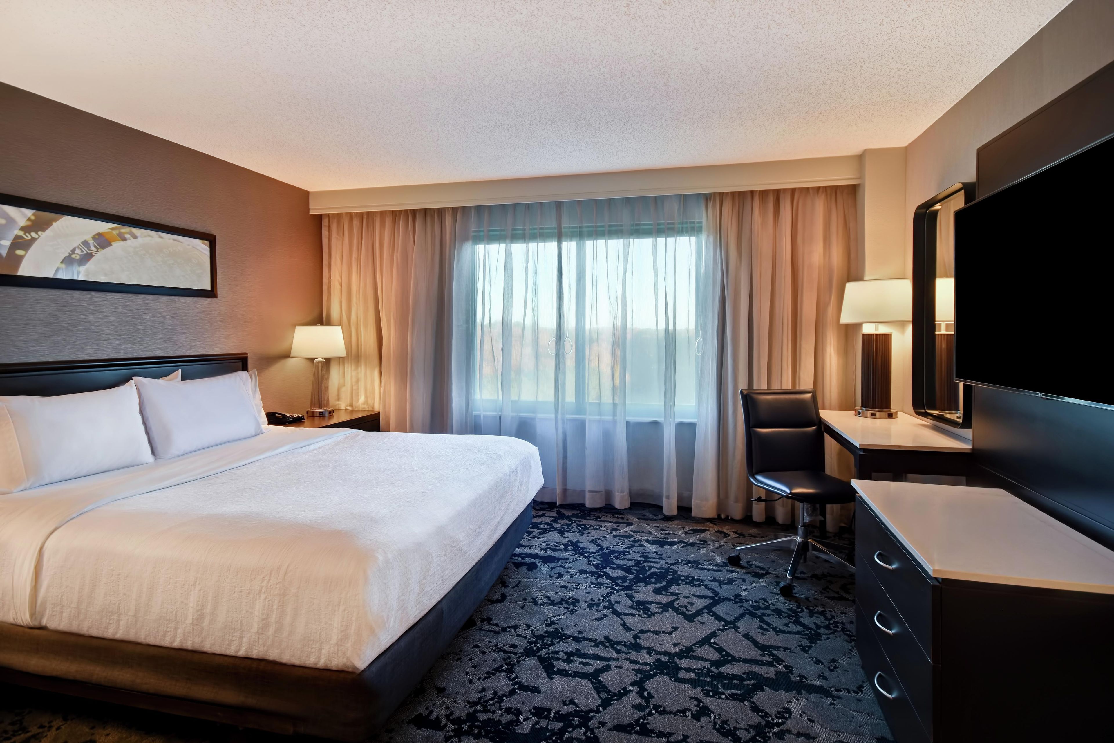 A hotel room with a bed, a desk, a chair, a TV, and a window with curtains.