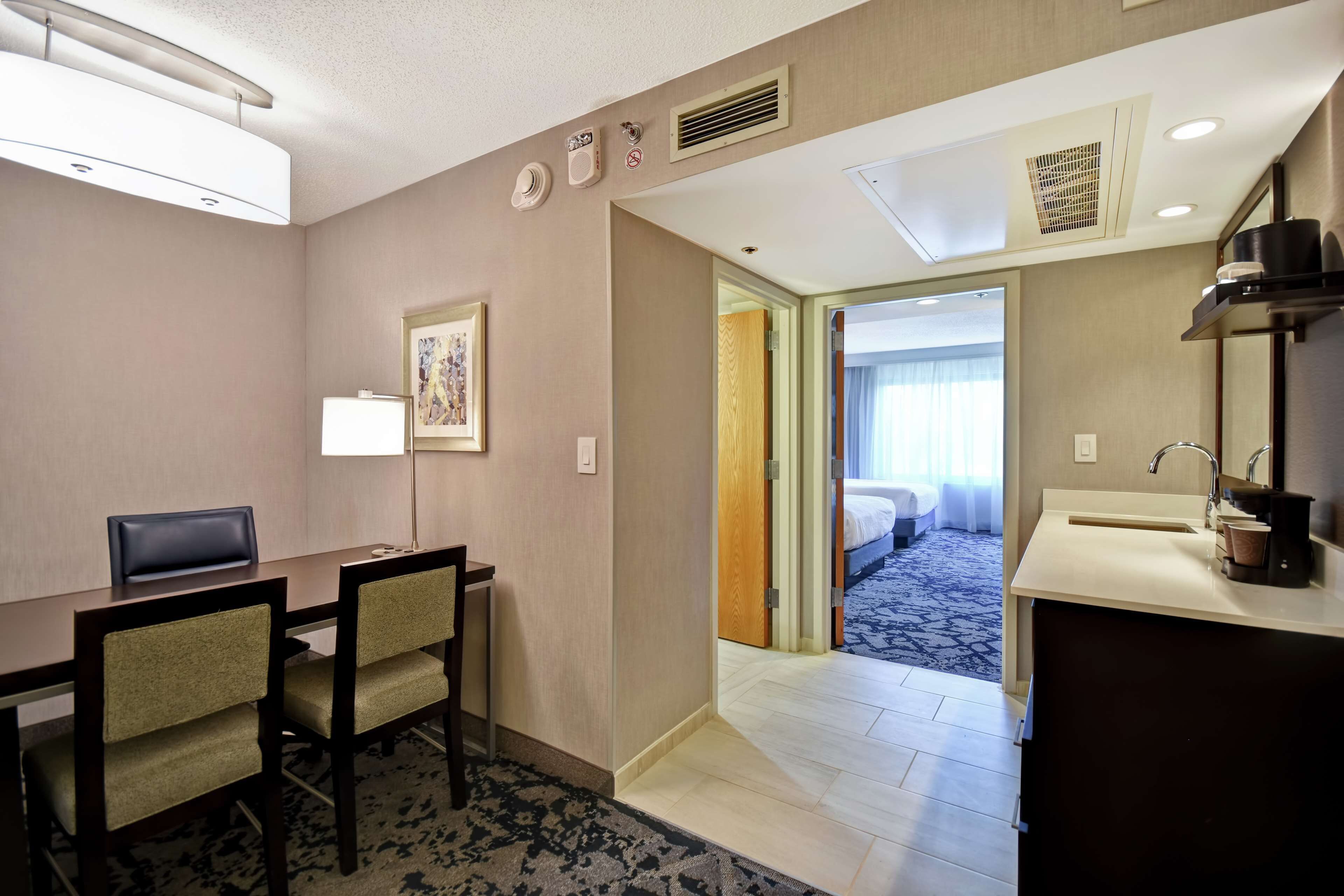 A hotel room with a dining area, a bed, a bathroom, and a view of the hallway.