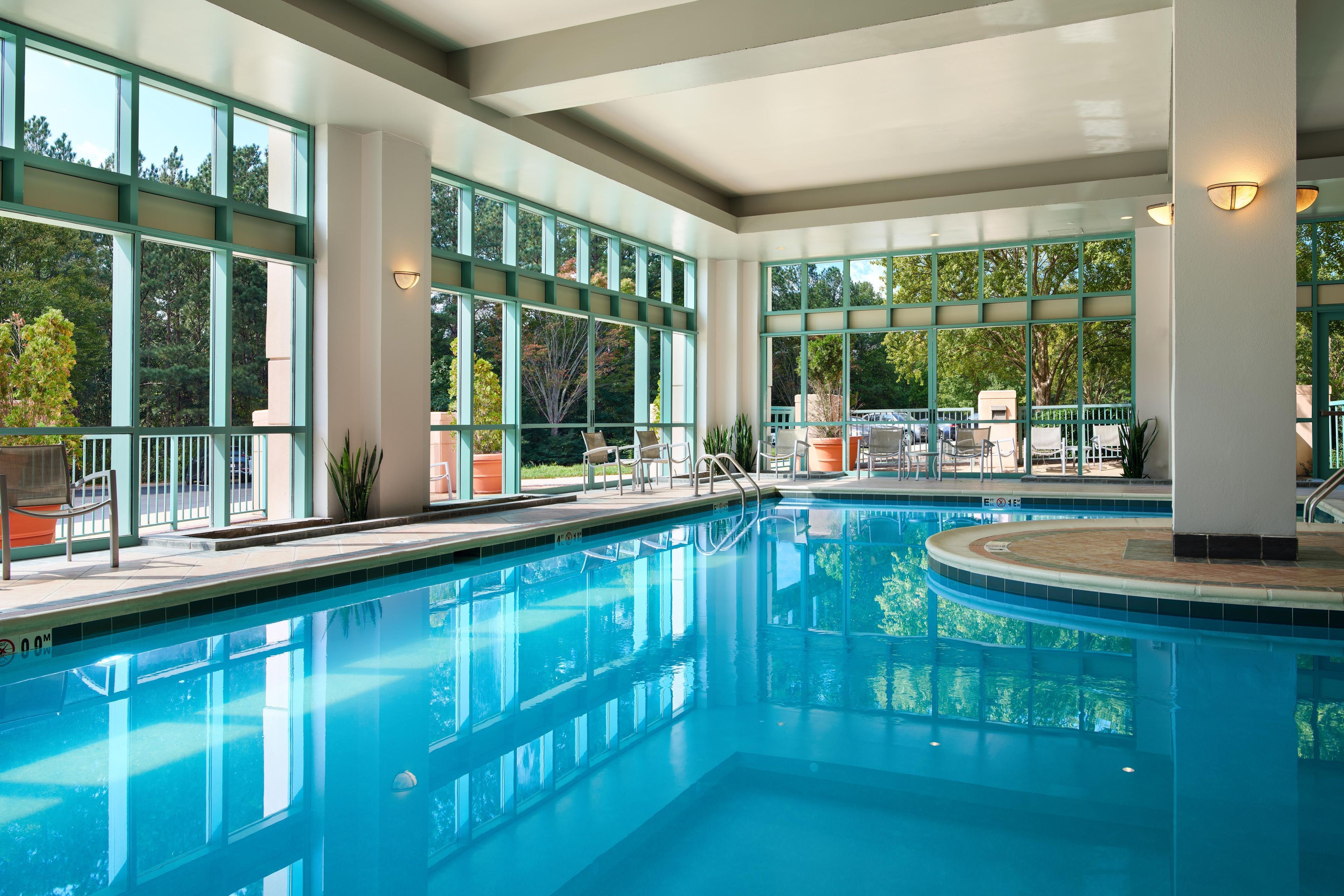 An indoor swimming pool with clear glass walls, multiple seating areas, and a view of the surrounding trees.
