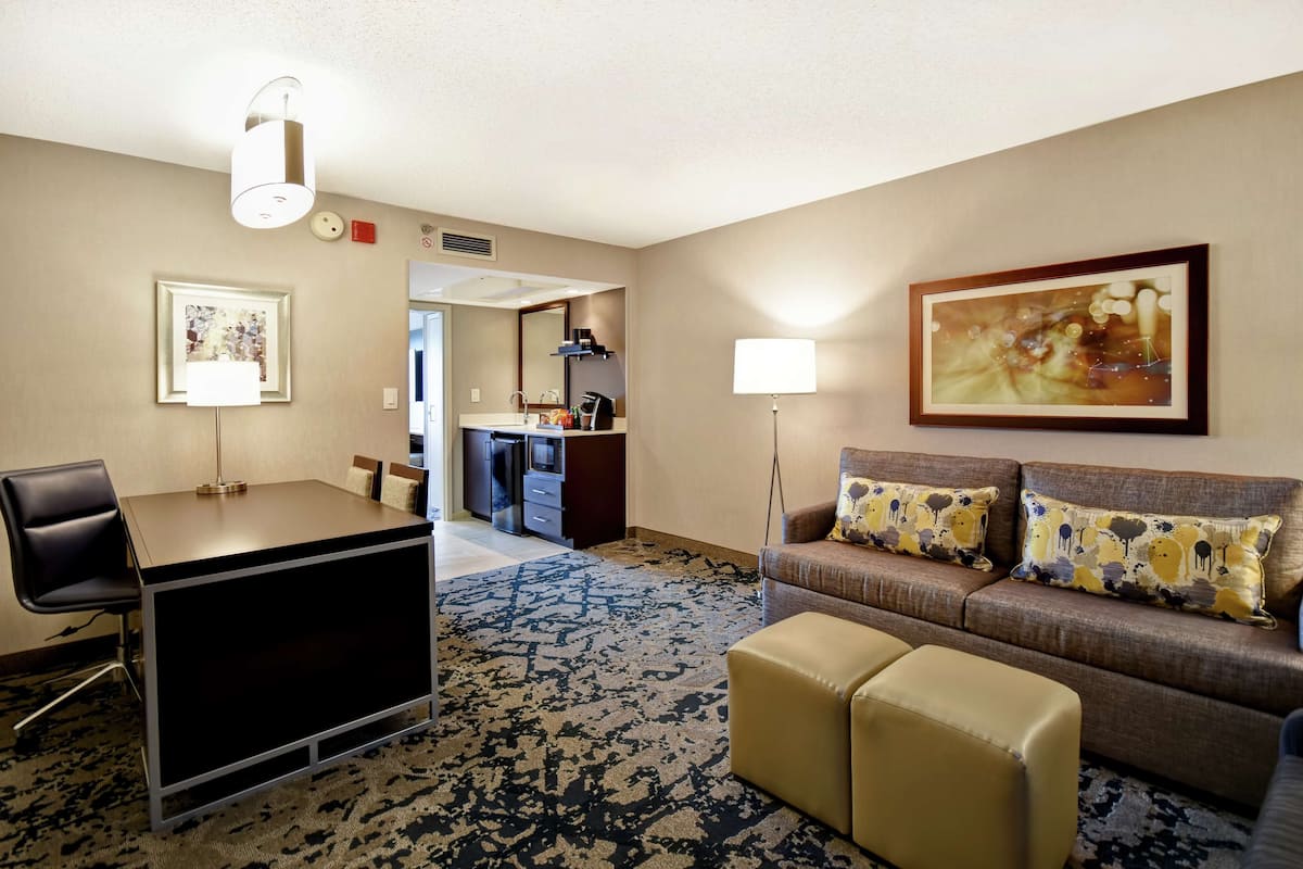 A hotel room with a desk, a sofa, a coffee table, a large painting, and a carpeted floor.