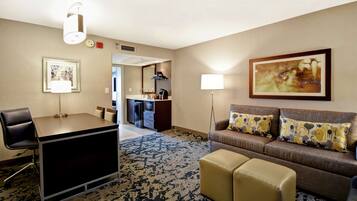 Habitación de hotel con escritorio, sofá, mesa de centro, un cuadro grande y piso alfombrado.
