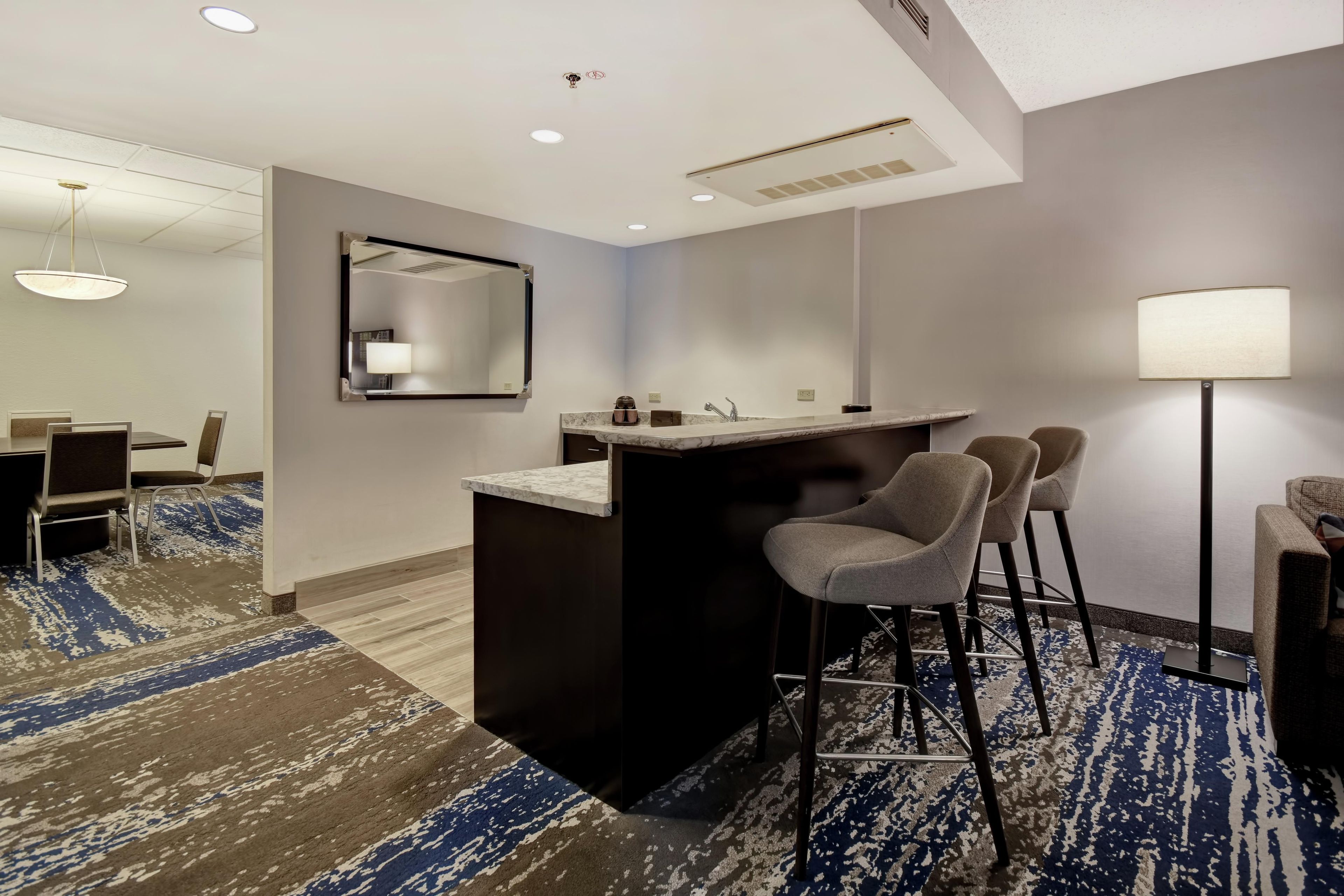 A hotel room with a bar area, a dining table with chairs, a large mirror, and a patterned carpet.