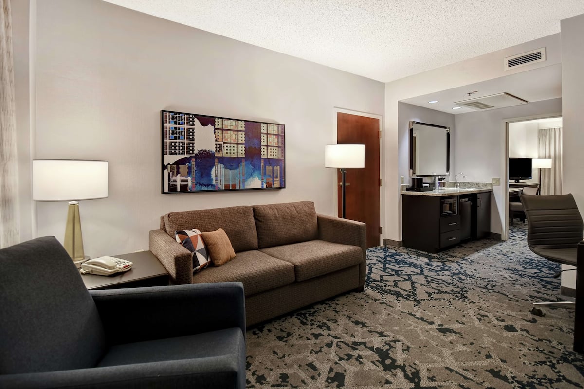 A hotel room with a sofa, armchair, desk, and a large painting on the wall.