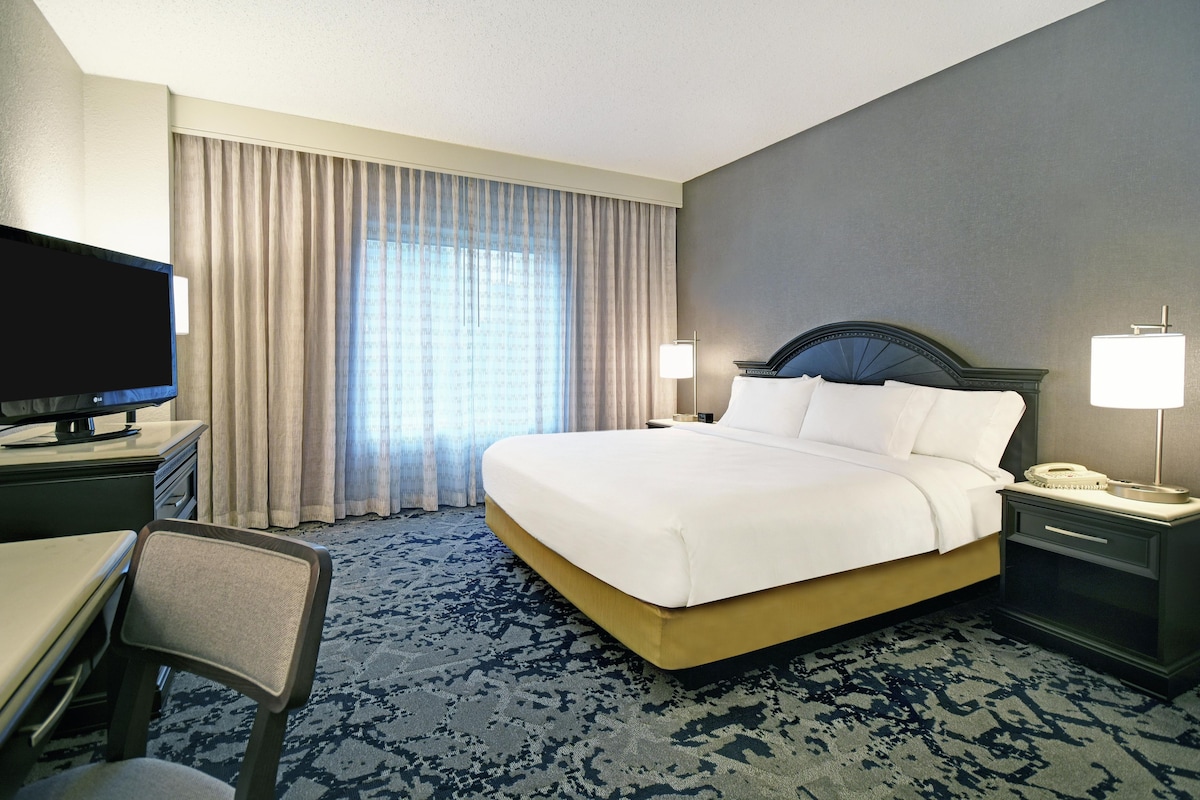 A hotel room with a bed, a desk with a television, a chair, a nightstand with a lamp, and a patterned carpet.