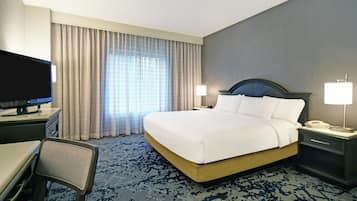 A hotel room with a bed, a desk with a television, a chair, a nightstand with a lamp, and a patterned carpet.