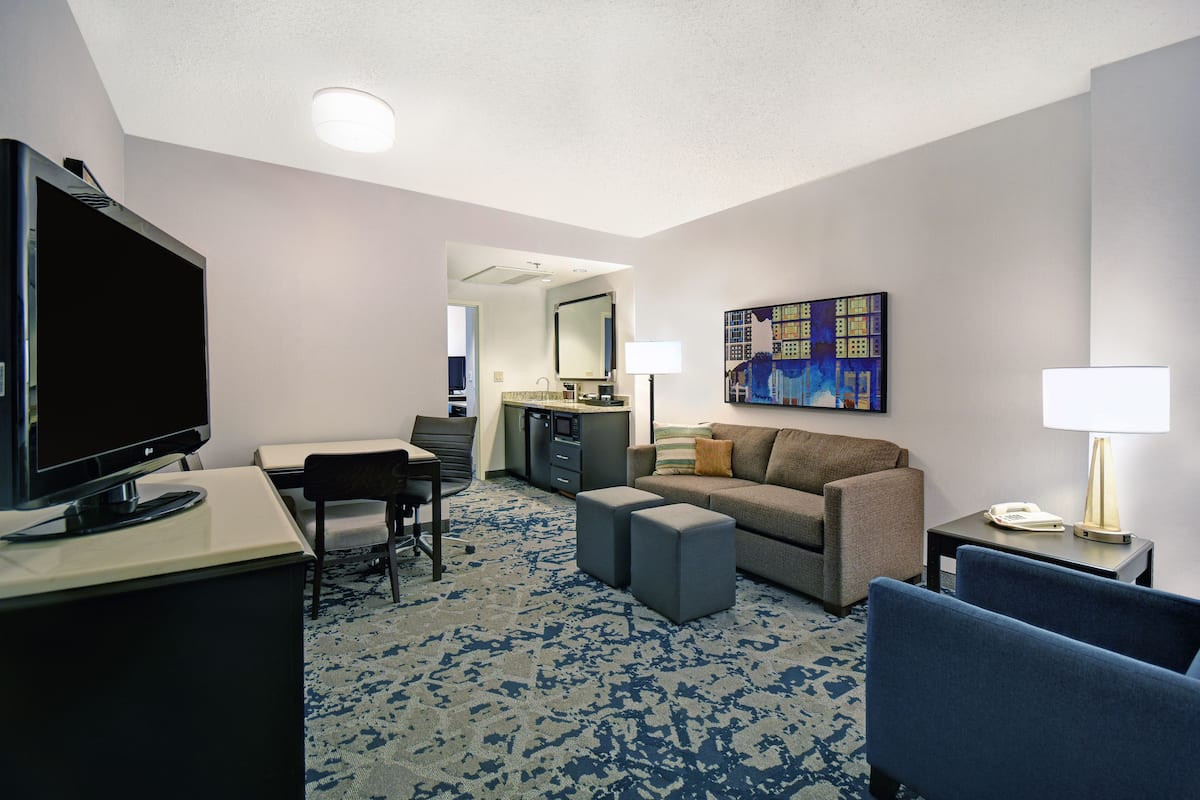 A hotel room with a flat-screen TV, a sofa, a desk, a chair, a small kitchenette, and a painting on the wall.