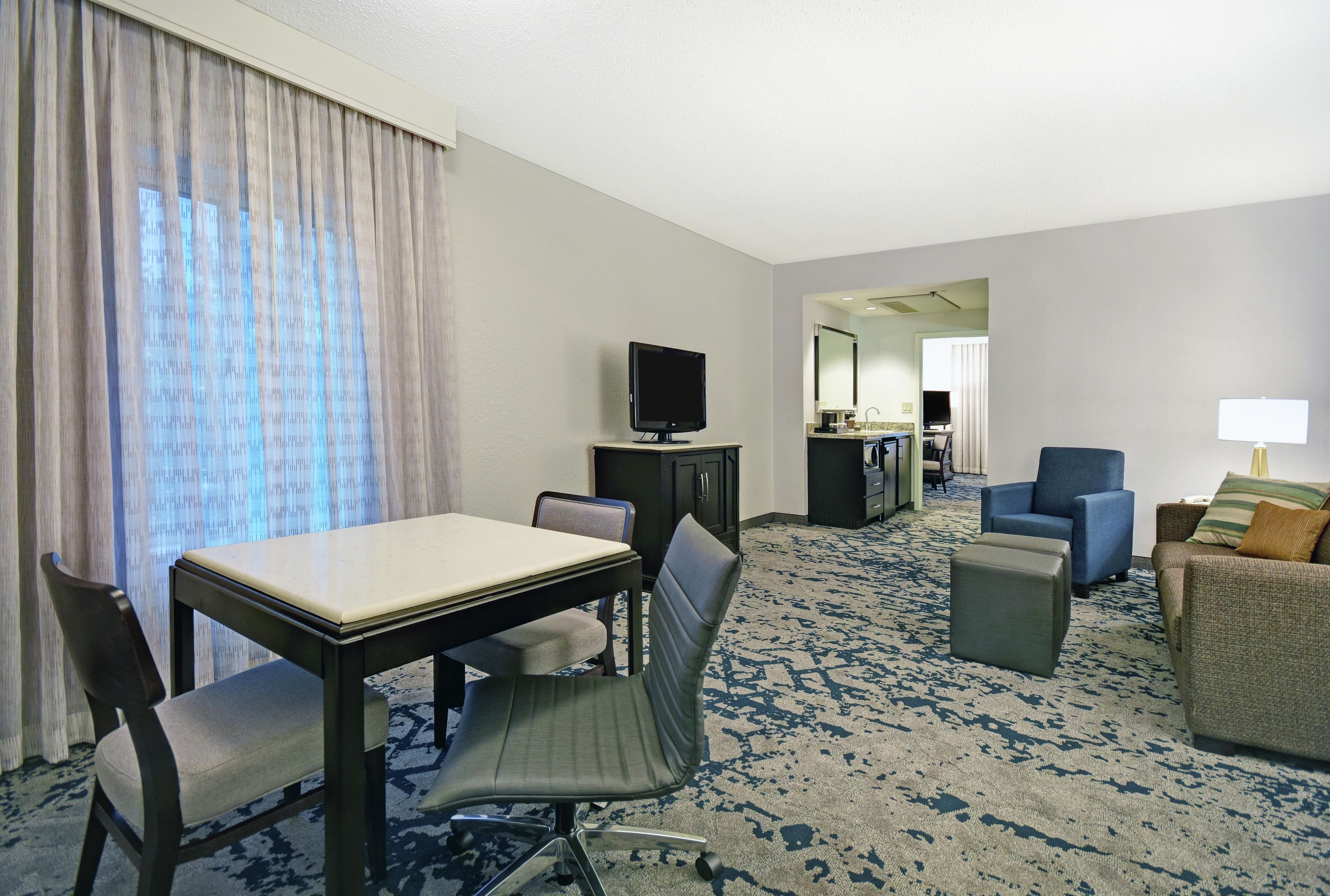 A hotel room with a dining table, two chairs, a television, a sofa, a small ottoman, and a lamp.