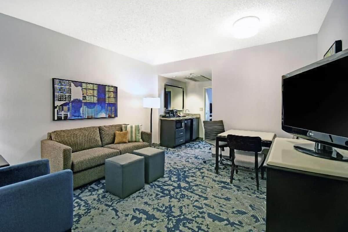 A hotel room with a sofa, ottoman, TV, desk, and a painting on the wall.