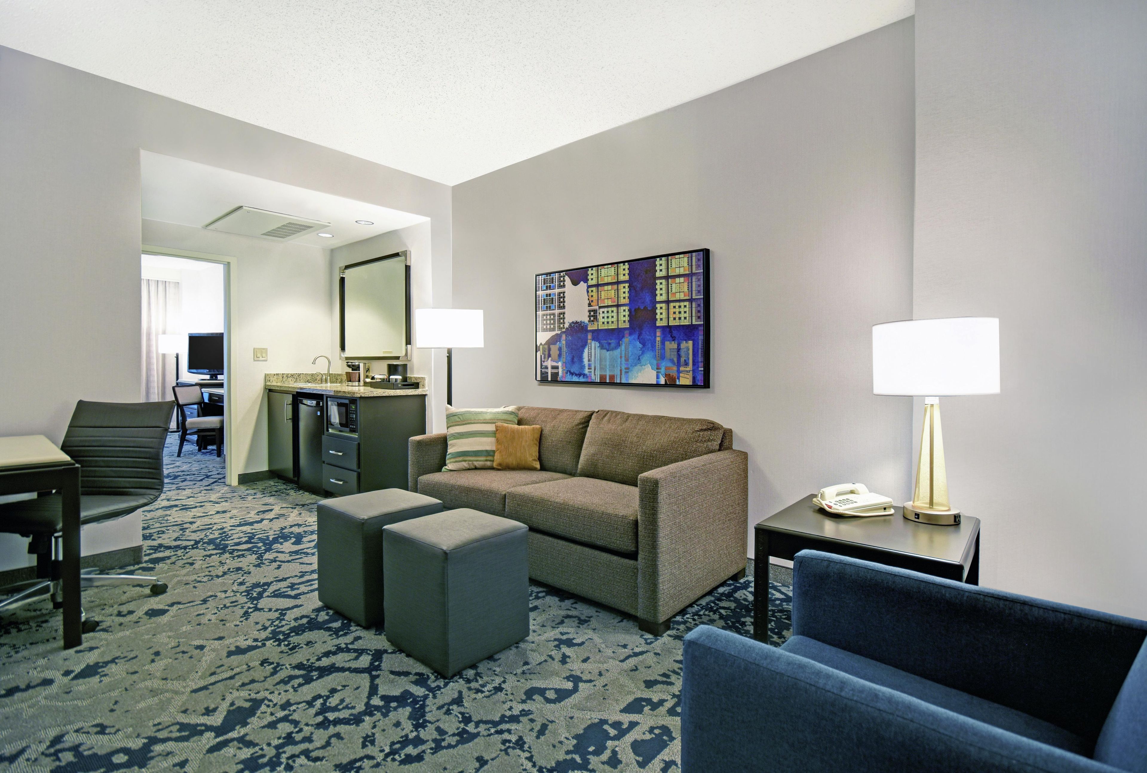 A hotel room with a sofa, armchair, ottoman, a desk with a chair, a small kitchenette, and a painting on the wall.