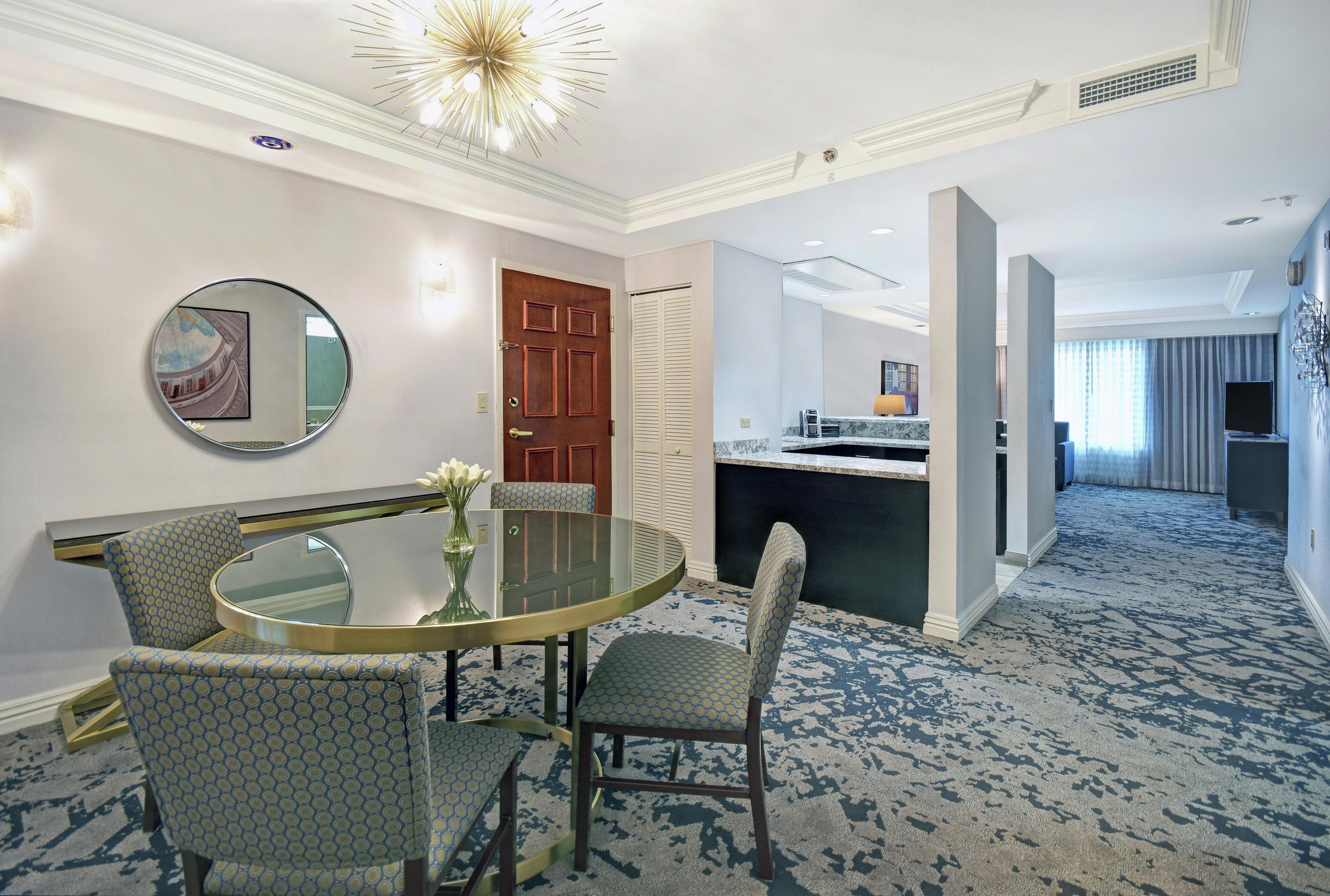 Habitación de hotel con una mesa de vidrio redonda, dos sillas con estampado, una puerta de madera, un espejo y vistas al pasillo.