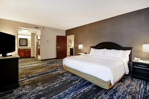 Habitación de hotel con una cama grande, televisor, escritorio con lámpara y baño visible a través de una puerta abierta.