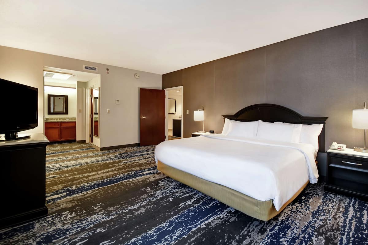 A hotel room with a large bed, a flat-screen TV, a desk with a lamp, and a bathroom visible through an open door.