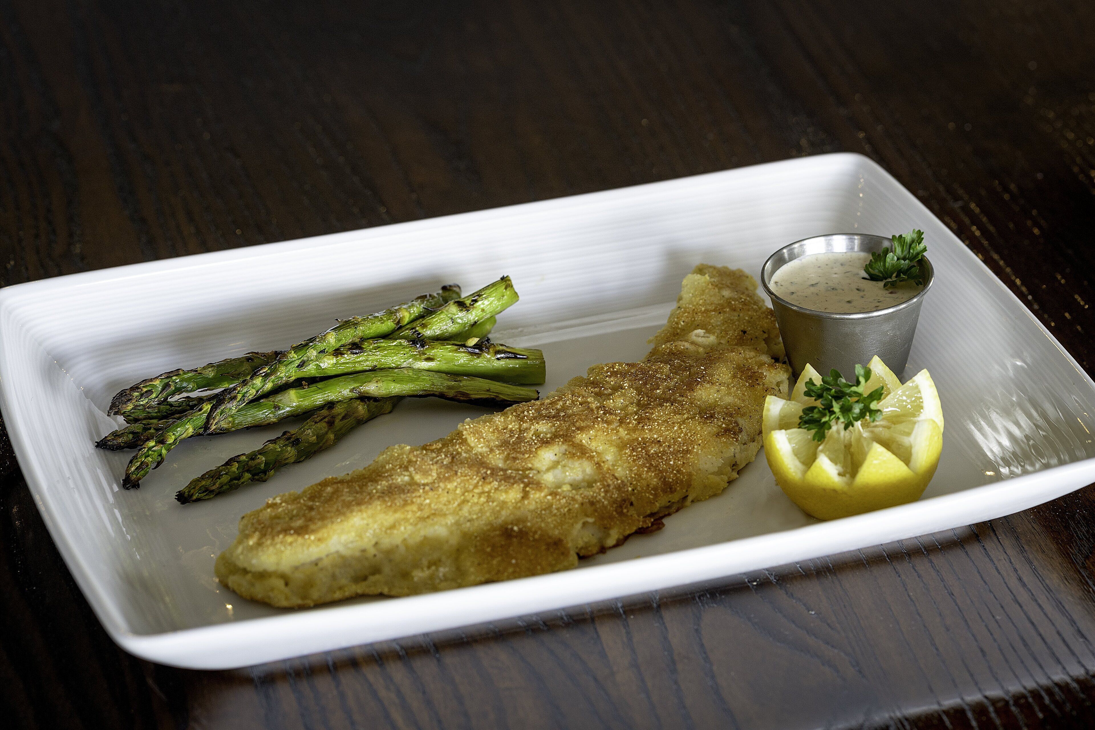 En vit, rektangulär tallrik med en panerad fiskfilé, sparris och en klyfta citron, serverad med en liten metallskål innehållande sås.