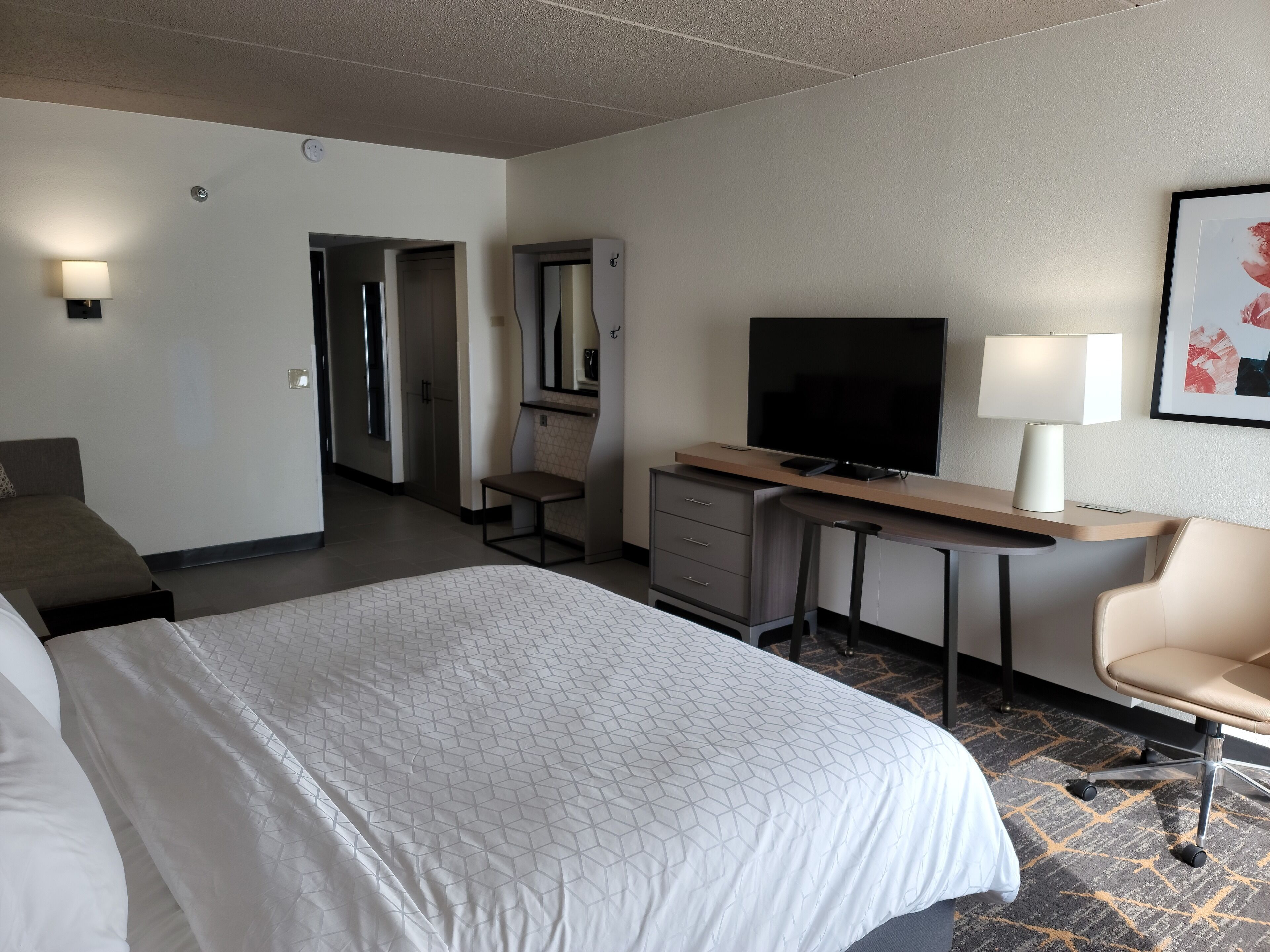 Habitación de hotel con una cama grande, un escritorio con televisor, una silla, una lámpara de escritorio y un cuadro en la pared.