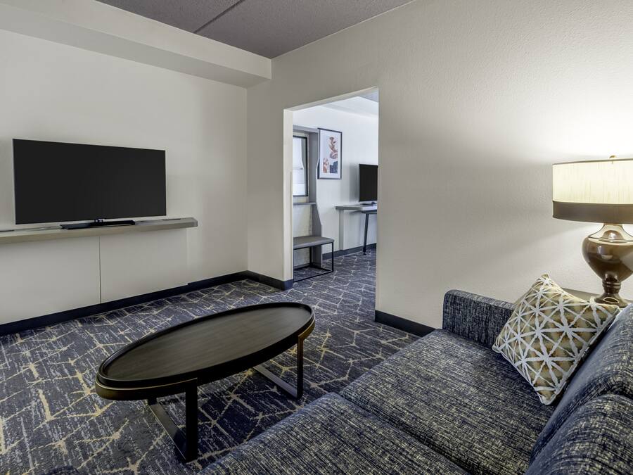 A hotel room with a flat-screen TV mounted on the wall, a sofa with a patterned cushion, a small table, and a lamp.