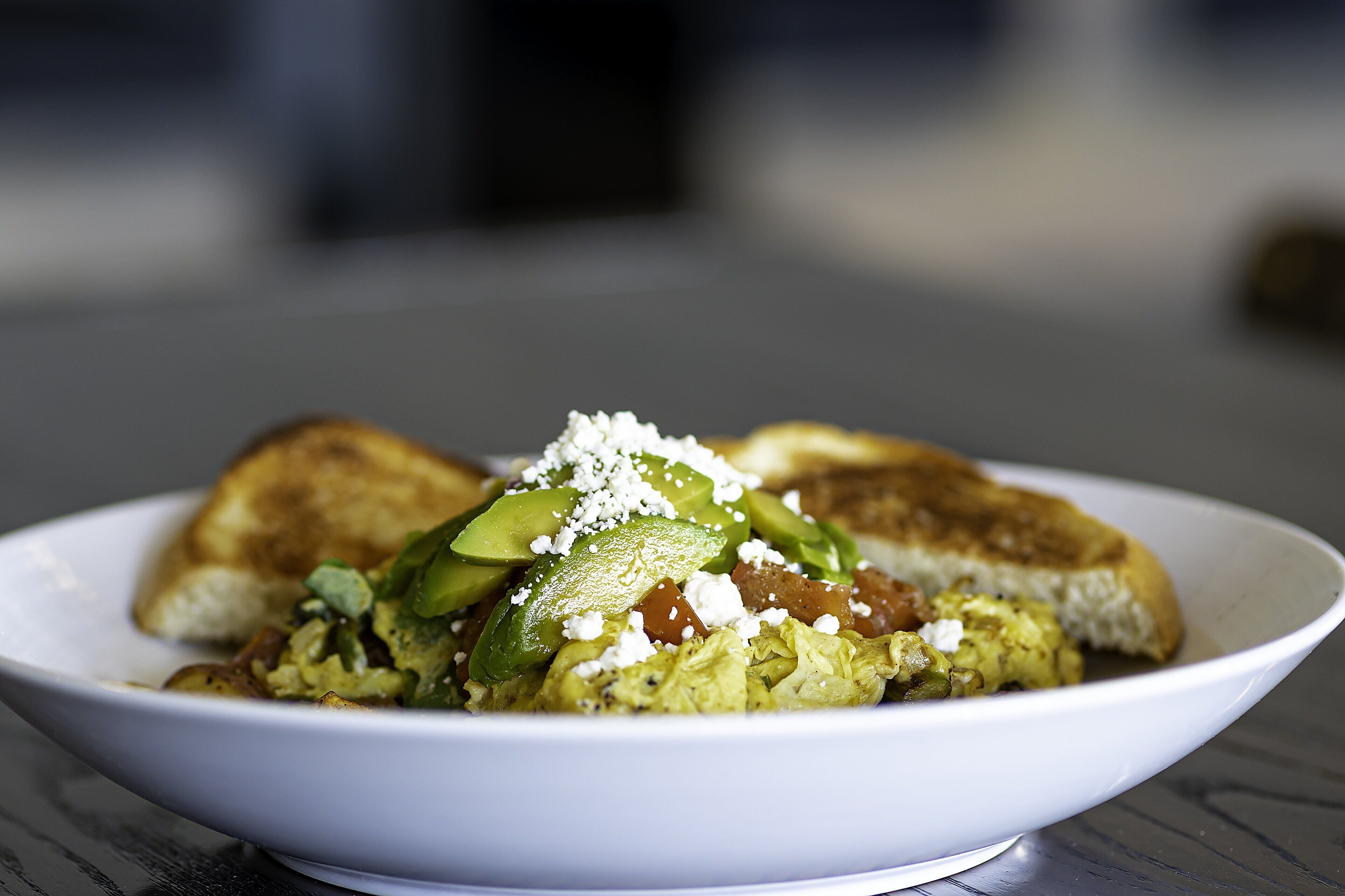 A white plate with a breakfast dish consisting of toast, avocado, and a green sauce, possibly containing eggs and greens.