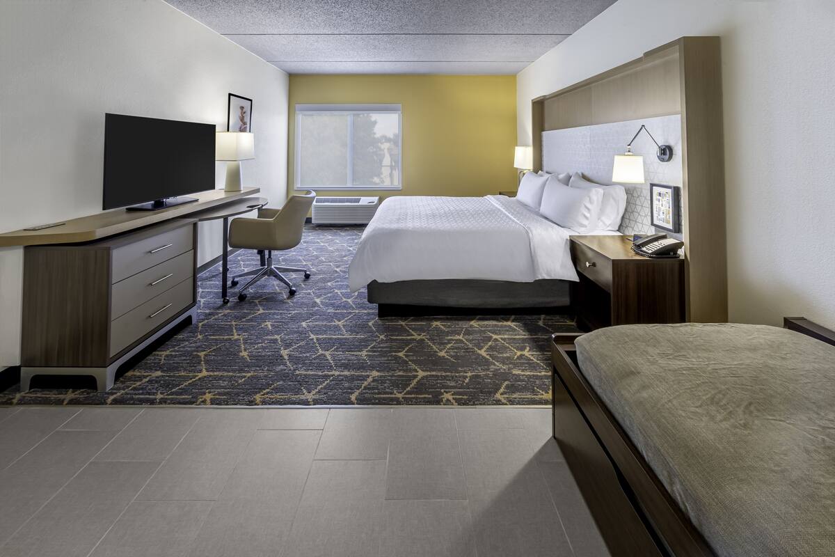 A hotel room with a bed, a desk with a TV, a chair, and a bedside table with a lamp.
