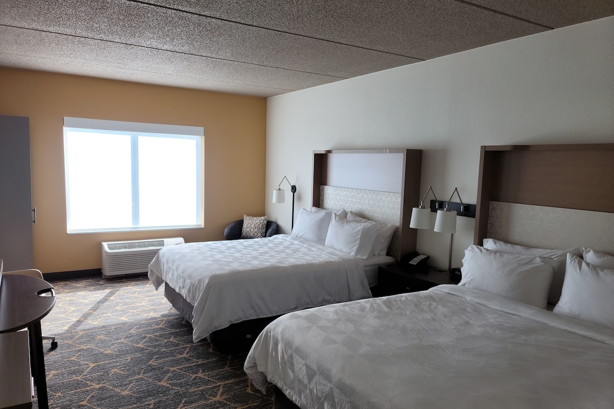 A hotel room with two beds, a desk, a chair, a window, and a wall-mounted lamp.