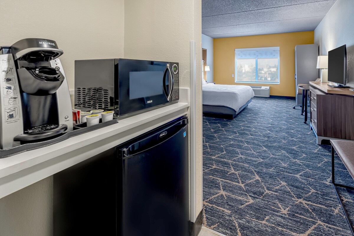 A hotel room with a kitchenette featuring a coffee maker, microwave, and refrigerator. There is a bed with a window, a desk with a lamp, and a TV.
