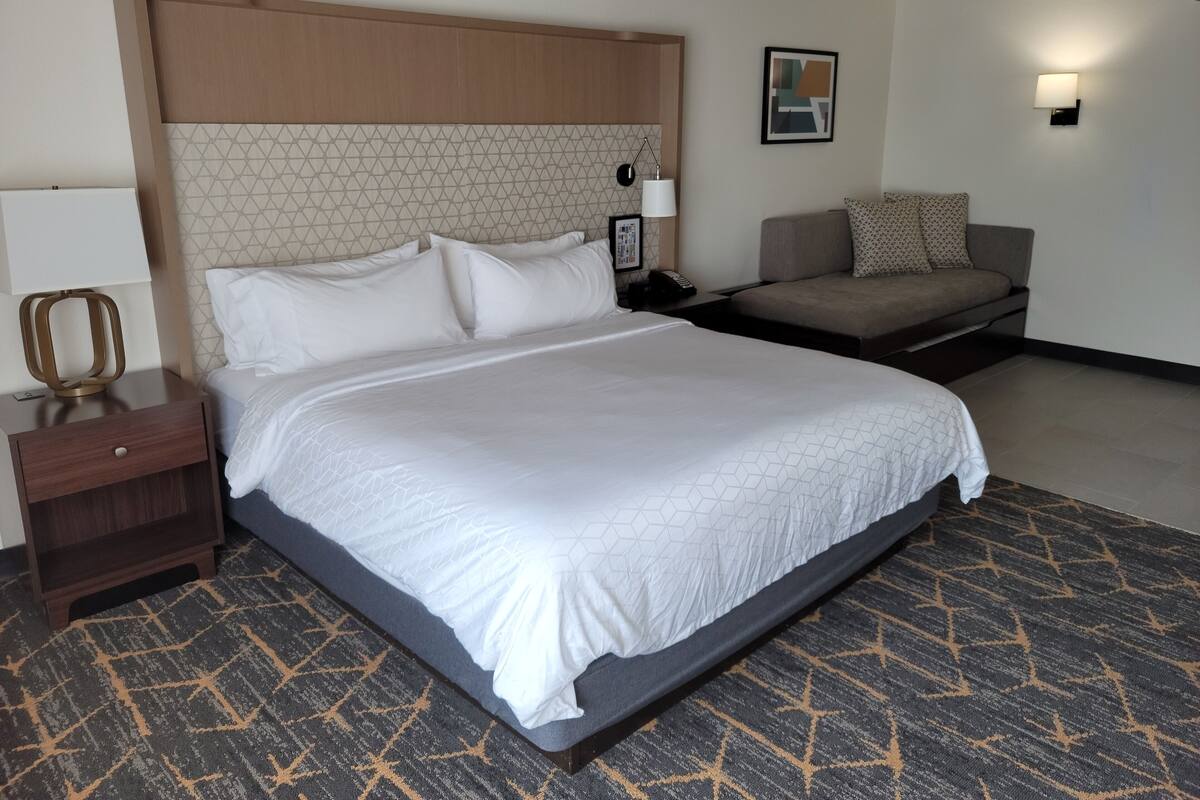 A hotel room with a large bed, a bedside table, a nightstand, a sofa, a wall-mounted lamp, and a framed picture on the wall.