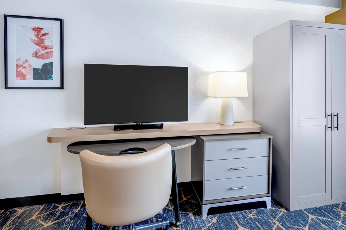A hotel room with a desk, chair, television, lamp, and wardrobe.