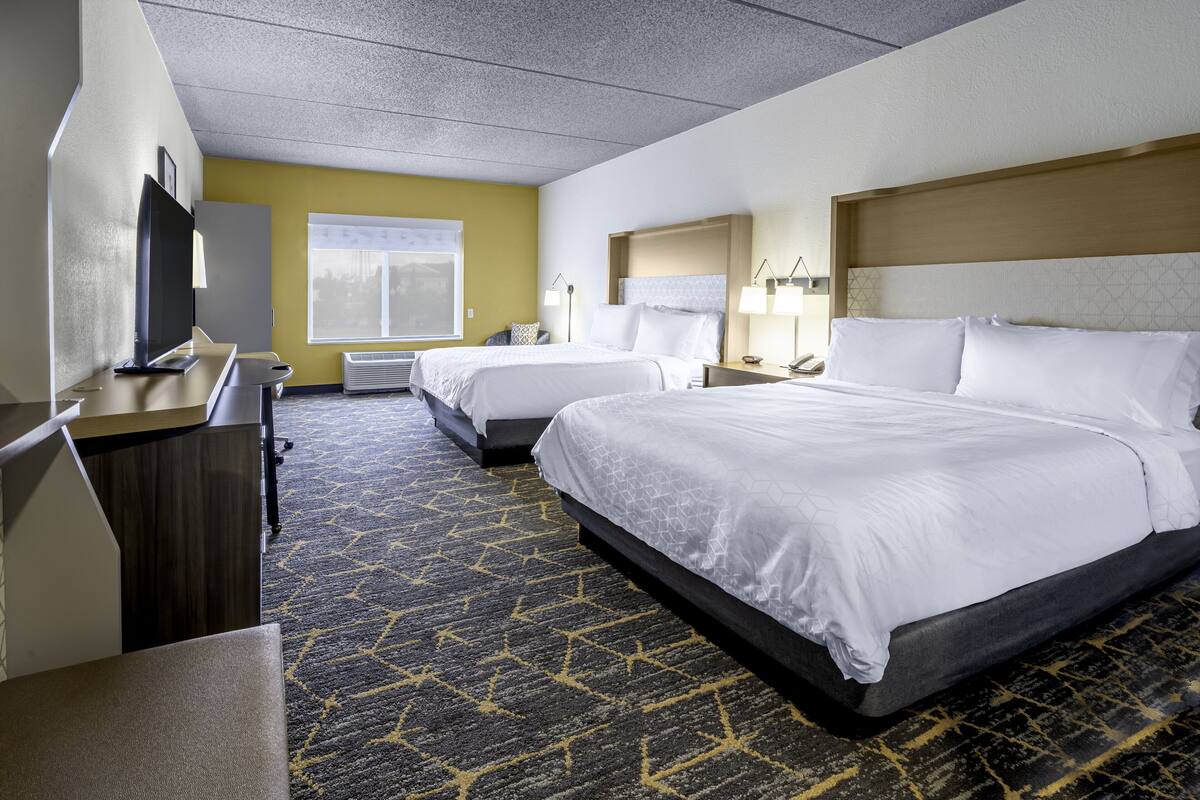 A hotel room with two beds, a desk with a flat-screen TV, a window, and a patterned carpet.