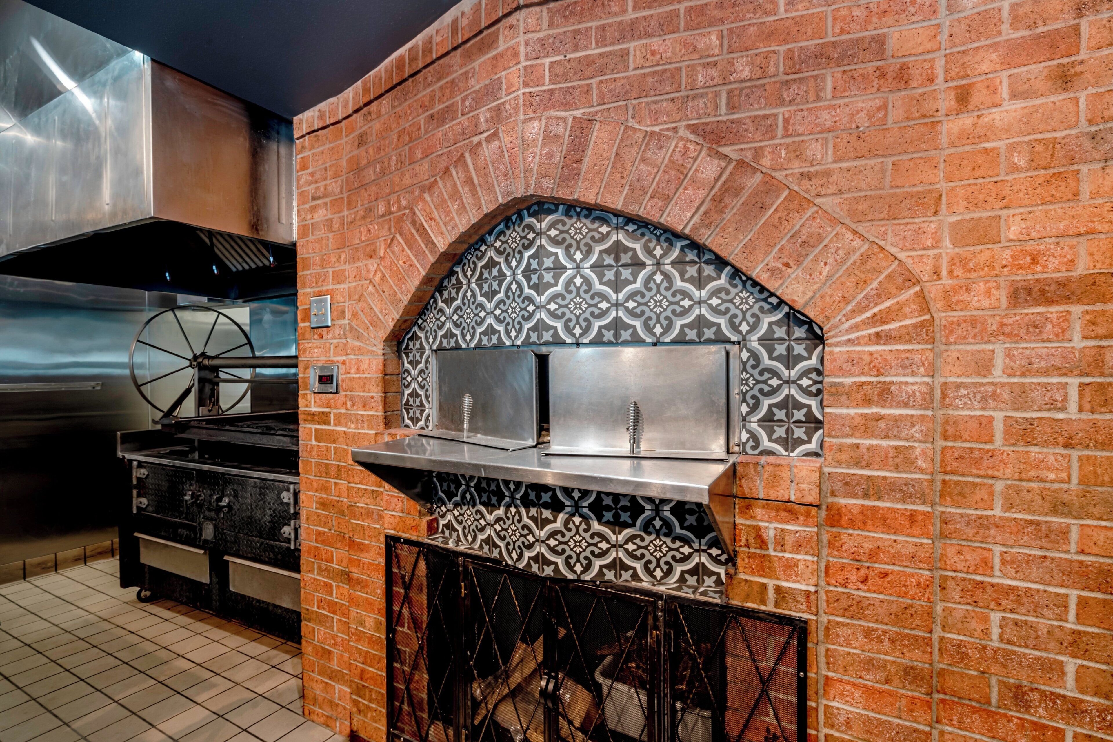 A brick oven with a decorative tile arch above it, a stainless steel grill, and a tiled floor.