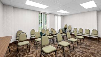 Una sala de conferencias con alfombra estampada, filas de sillas, una mesa y un ventanal con persianas verticales.