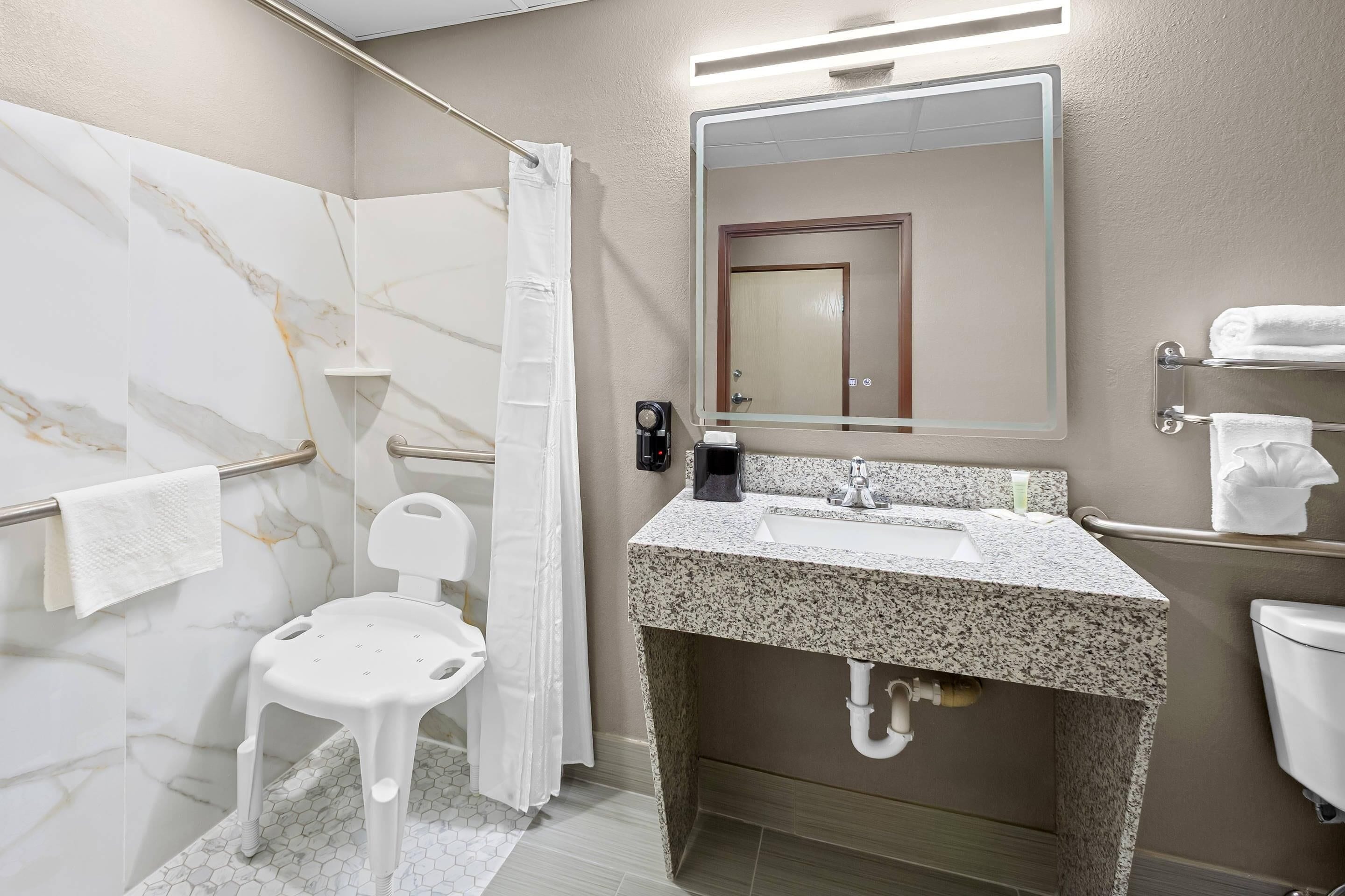 Un baño con pared de mármol, ducha con silla blanca, lavabo con espejo y un inodoro.