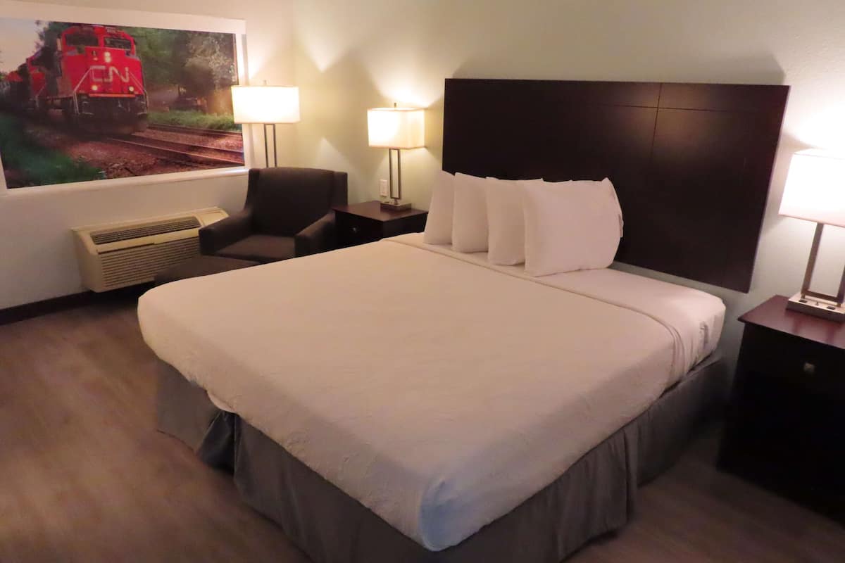 Ein Hotelzimmer mit einem großen Bett, zwei Nachttischlampen, einem Sessel, einem Heizgerät und einem an der Wand montierten Fernseher, der einen Zug zeigt.