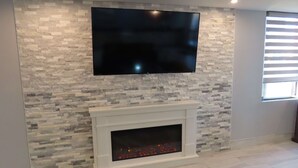 A living room with a stone-patterned wall, a flat-screen TV mounted above a fireplace, and a window with blinds.