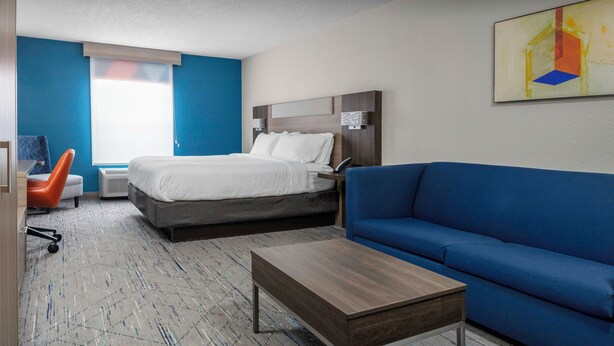 A hotel room with a bed, a blue sofa, a wooden coffee table, a desk with a chair, and a window with blinds. - Holiday Inn Express & Suites Kimball by IHG (South Pittsburg)