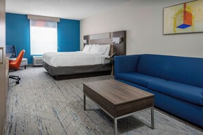 A hotel room with a bed, a blue sofa, a wooden coffee table, a desk with a chair, and a window with blinds. - Holiday Inn Express & Suites Kimball by IHG (South Pittsburg)