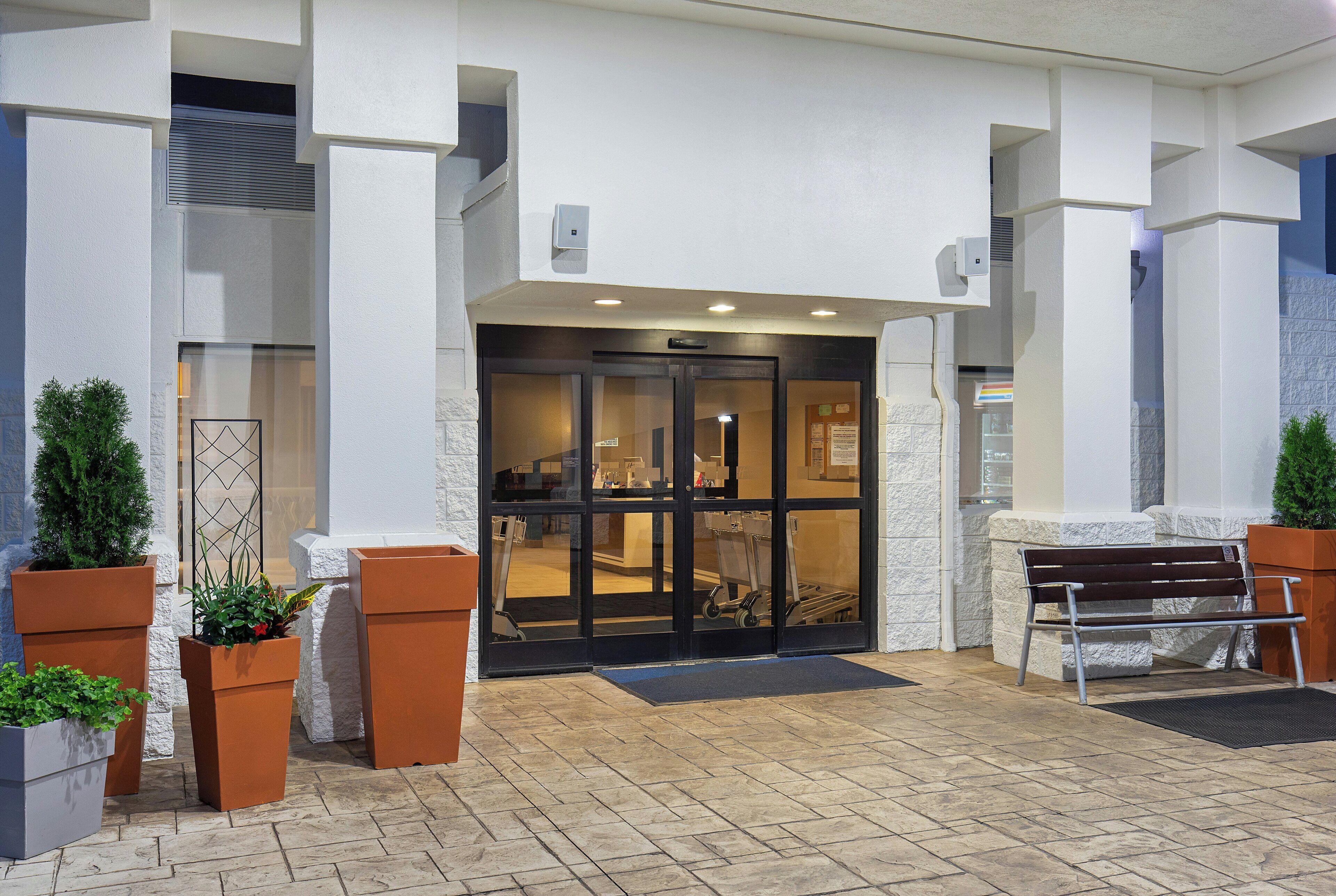 A hotel entrance with a glass door, potted plants, a bench, and a tiled pavement.