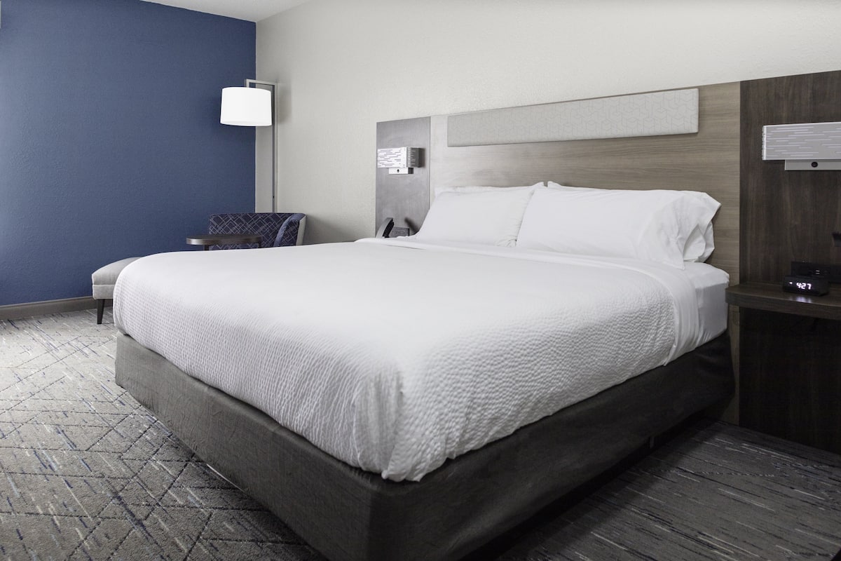 A modern hotel room with a large bed, a bedside table, a chair, and a wall-mounted lamp.