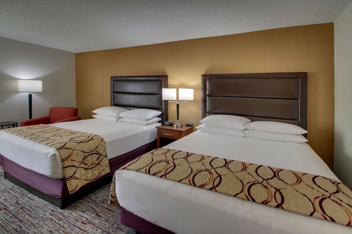 A hotel room with two beds, a red chair, a nightstand with a lamp, and a carpeted floor.