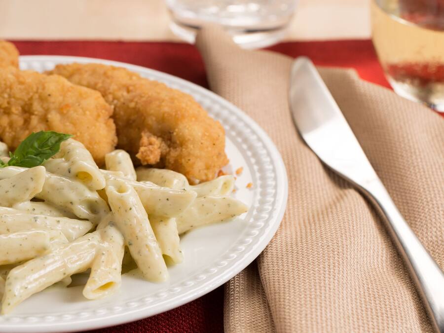 Een bord pasta met gepaneerde schnitzels en garnering, met een vork en mes op een tafelkleed.