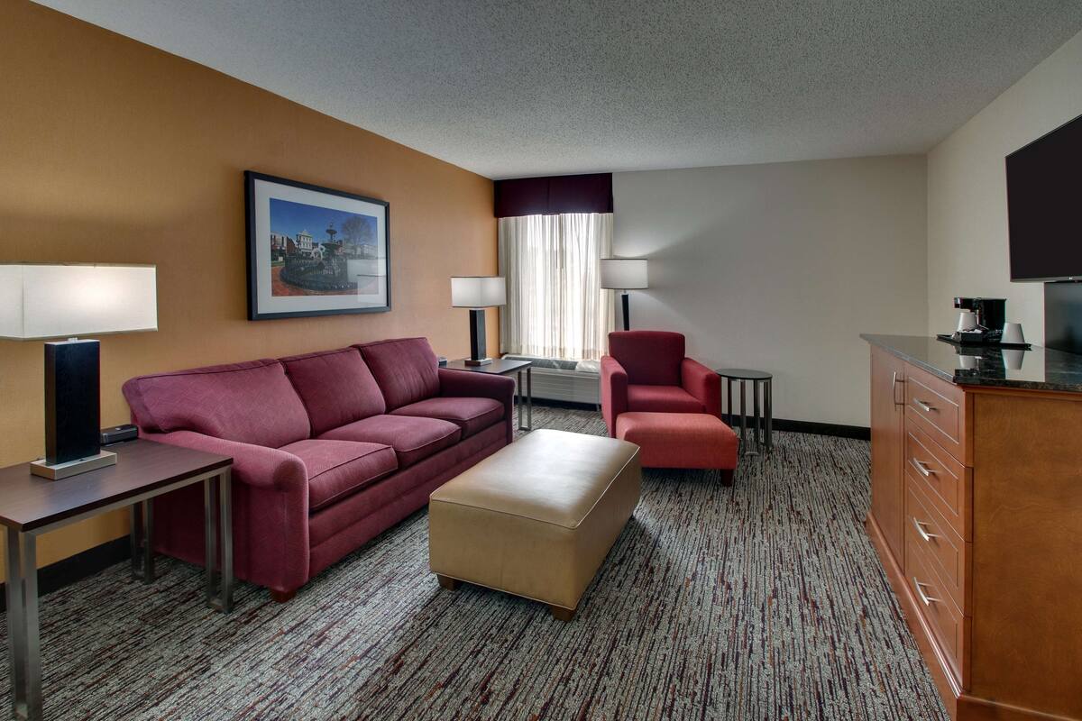 A hotel room with a red sofa, a beige ottoman, a red armchair, a small table, a lamp, a TV, and a cabinet.