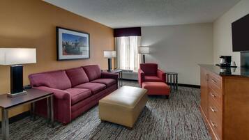 Una habitación de hotel con un sofá rojo, un reposapiés beige, un sillón rojo, una mesita, una lámpara, un televisor y un armario.