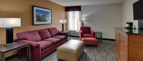 A hotel room with a red sofa, a beige ottoman, a red armchair, a small table, a lamp, a TV, and a cabinet.