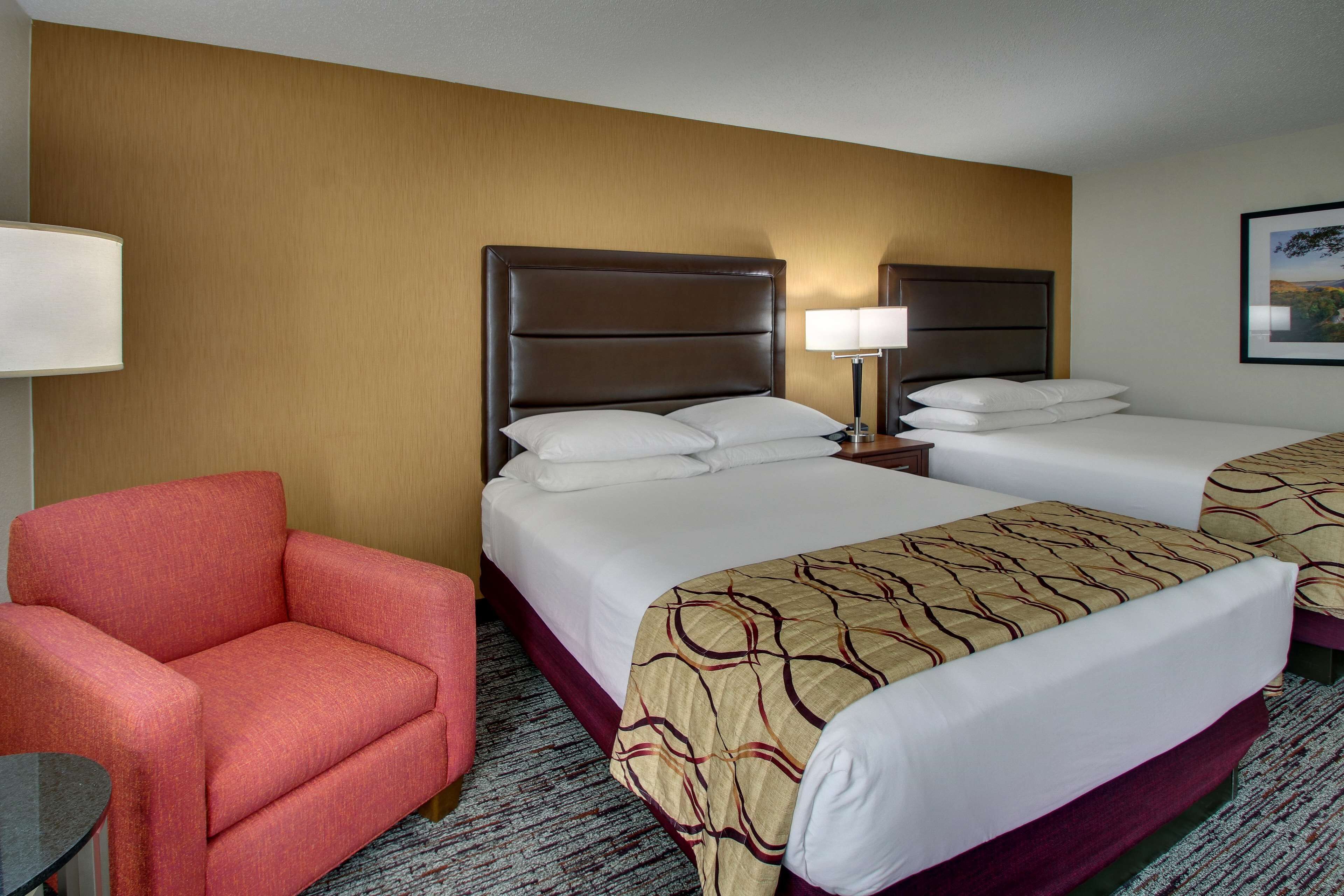 A hotel room with two beds, a red armchair, a small table, and a lamp.