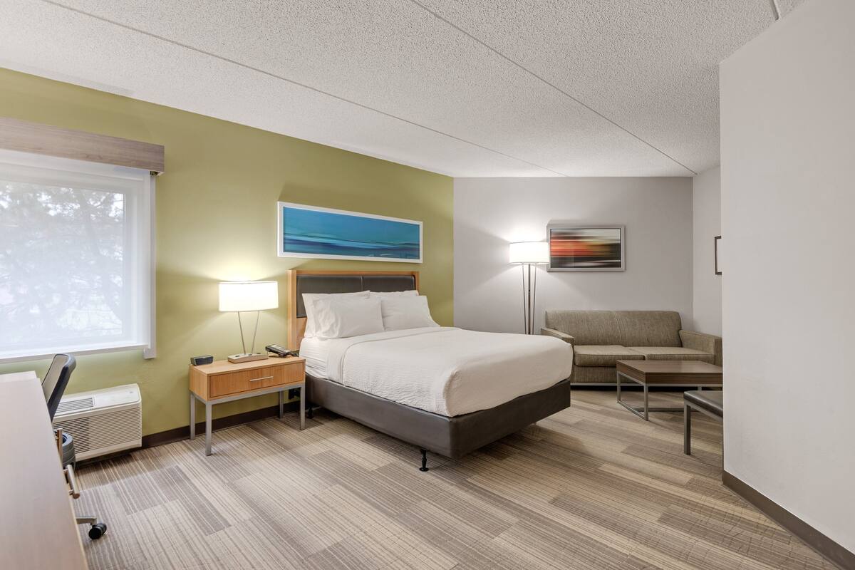 A hotel room with a bed, a desk, a chair, a lamp, and a painting on the wall.