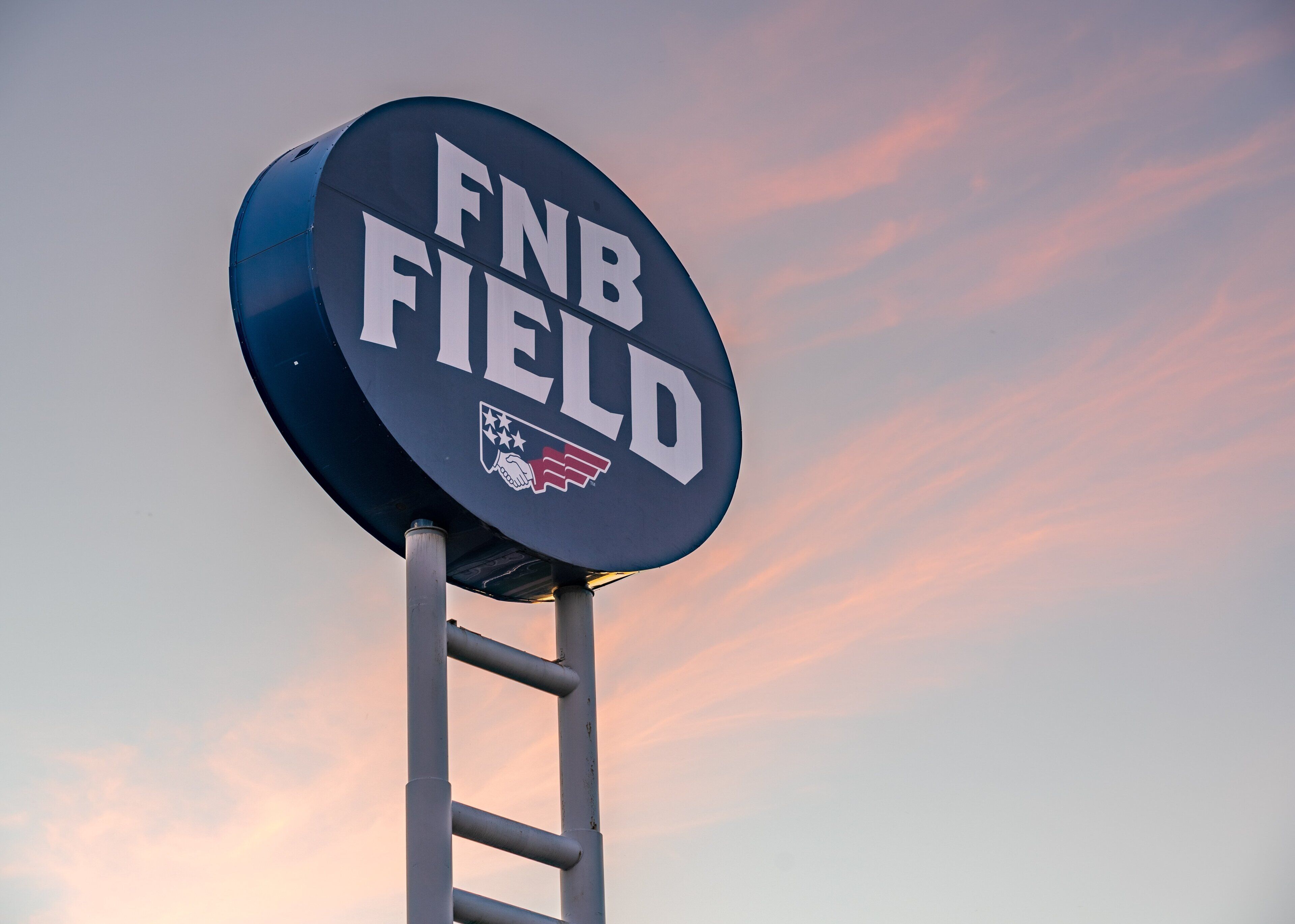 Uma placa com o texto 'FNB FIELD' e um logotipo, fixada em um poste de metal contra um céu com nuvens.
